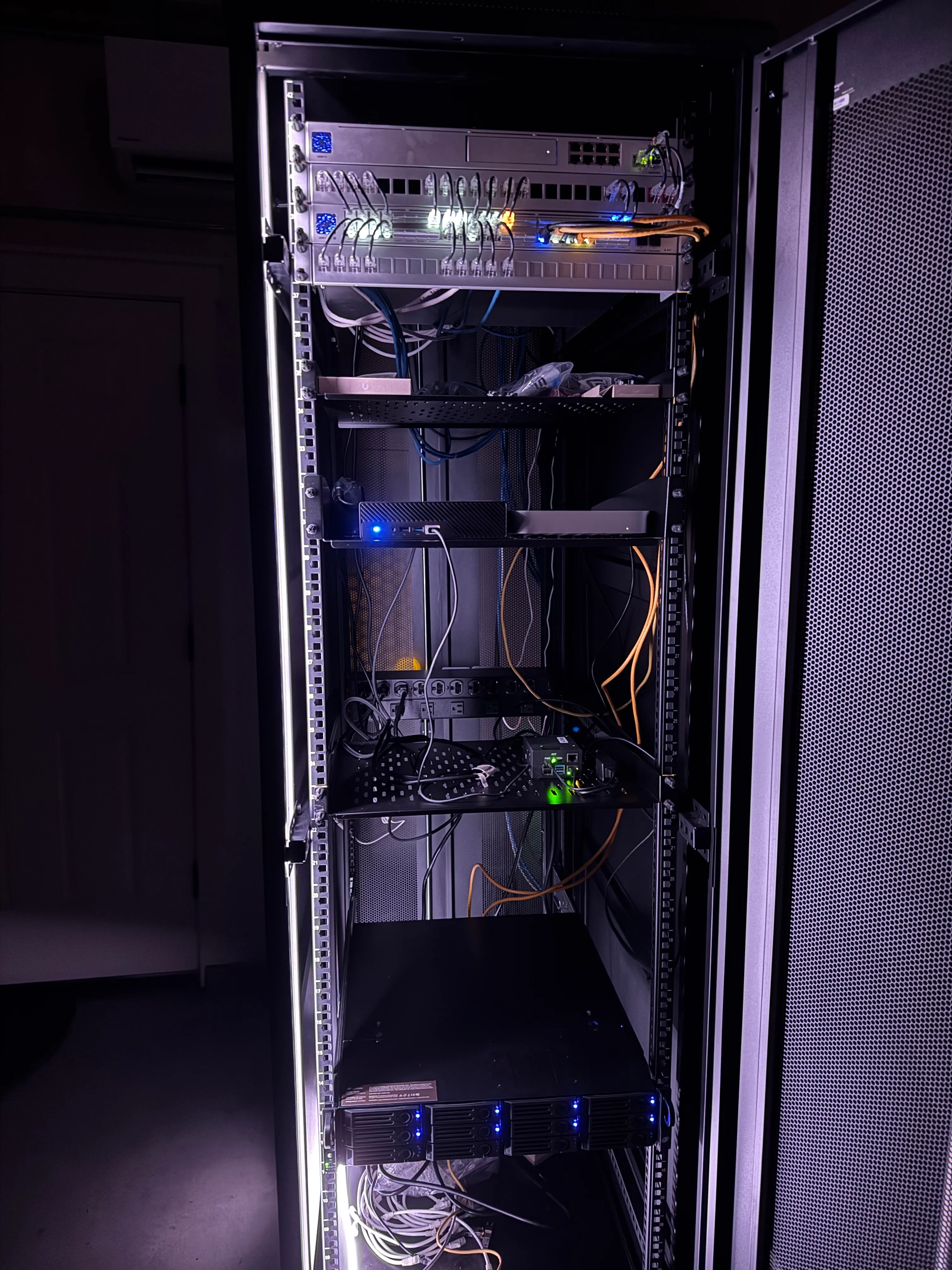 A 42U server rack in the dark. At the top is a UDM Pro and a USW Pro XG 48 PoE switch surrounded by keystone ports. Then a shelf with miscellanea, a shelf with a Mini PC and Mac mini, and a shelf with a Pi-KVM. Near the bottom is a 12-bay TrueNAS Mini R. The photo is brightly illuminated by blinkenlights from the equipment and a violet-tinted glow from an LED light strip embedded in the edge of the rack.
